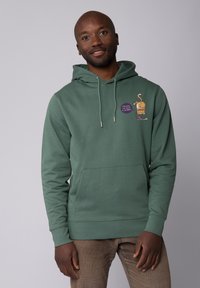 Groene hoodie gemaakt van zacht materiaal, met een voorzak, capuchon met trekkoord en een speelse graphic van een hond met een paarse label.