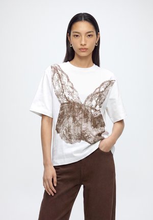 Femme aux cheveux noirs et lisses portant des boucles d'oreilles créoles dorées, un t-shirt blanc avec un grand imprimé papillon marron, et un pantalon marron.