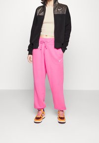 Svart zip-up jacka med glittriga axelpaneler, kombinerad med klara rosa sweatpants. Outfiten avslutas med bruna och orange sneakers.