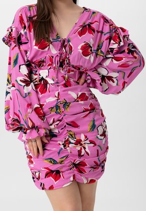 Robe fleurie violette avec manches longues bouffantes, devant froncé et col en V noué à la poitrine, portée par une personne avec une main sur la hanche.