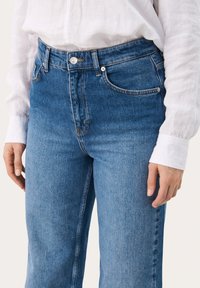 Jeans de tiro alto de color azul en un lavado medio con una textura suave. Presenta botón metálico, bolsillos laterales y detalles de costura sutiles.