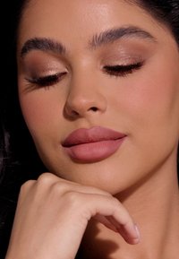 Close-up van een gezicht met matte mauve lippen, licht bronzen oogschaduw, gedefinieerde wenkbrauwen en een egale huid. Zachte verlichting accentueert de kenmerken.