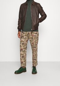 Man som bär en brun läderjacka, mörkgrön polotröja, kamouflagemönstrade byxor och gröna snörskor, står med ena handen i fickan.