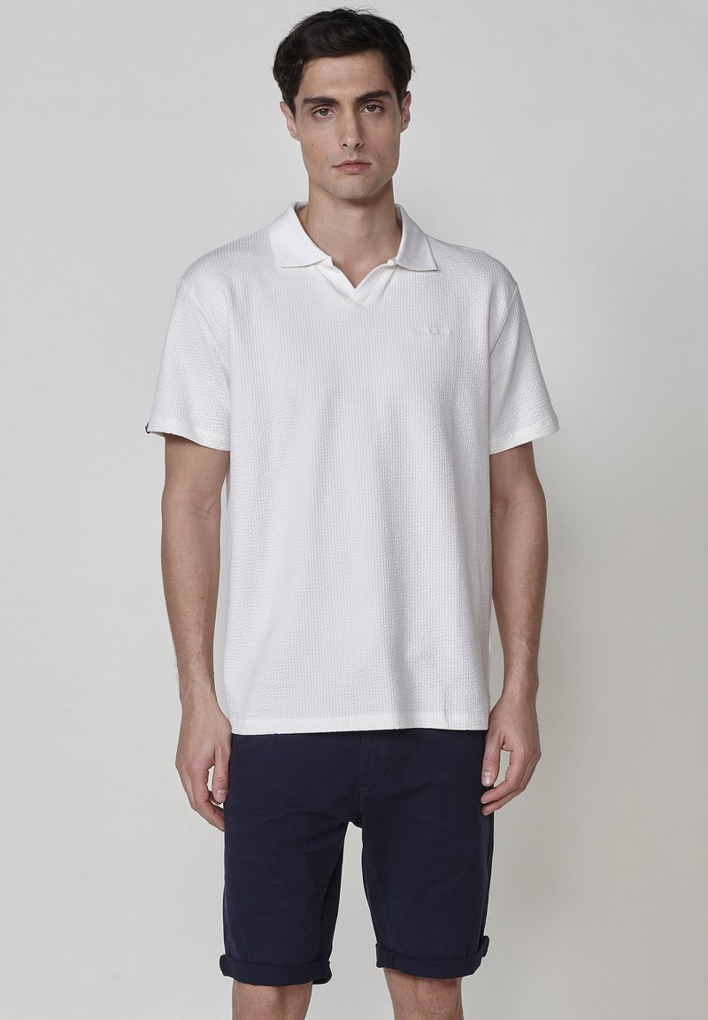 Vit kortärmad polo med texturerad tyg, V-ringning och uppvikta ärmar, i kombination med marinblå shorts.