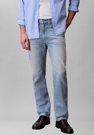 Mann trägt hellblaue Jeans, schwarze Lederschuhe, weißes T-Shirt und ein blau-weiß gestreiftes Hemd mit hochgekrempelten Ärmeln.