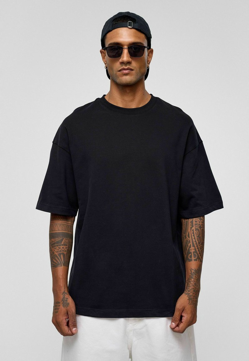 Zwarte oversized t-shirt van katoen, met korte mouwen en een ronde hals. Model draagt een zonnebril en een zwarte pet. Lichte achtergrond.