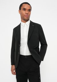 Sort blazer med slim fit design, der har to knapper og en brystlomme. Bærert over en hvid skjorte med krave, glat tekstur.