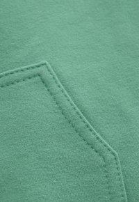 Close-up van mintgroene stof met gedetailleerde stiksels langs de rand van een rechthoekige zak.