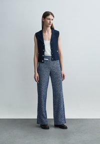 Kobieta ubrana w granatową bezrękawnikową kamizelkę na guziki, biały top, niebieskie szerokie spodnie o fakturze i czarne buty, stojąca na tle jasnego, jednolitego tła.