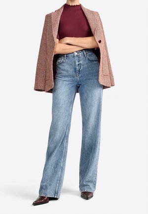 Personne portant un haut bordeaux, un jean large taille haute bleu clair, des chaussures pointues marron et un blazer à motifs posé sur les épaules.