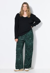 Femme aux cheveux ondulés portant un pull noir, un pantalon large en paisley vert et des baskets blanches, se tenant avec une main sur la hanche.