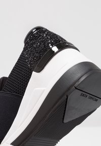 Chaussure de sport noire avec une tige en mesh texturé, un accent noir brillant et une semelle blanche avec détails rainurés. Surcouche scintillante au talon.