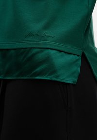 Zielony top sportowy wykonany z miękkiego materiału, charakteryzujący się warstwowym designem z satynowym wykończeniem na dole oraz subtelnym detalem z logo.