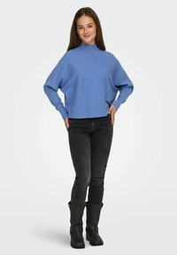 Mlada žena stoji s rukama na bokovima, wearing plavi pulover, crne traperice i crne čizme uz jednostavnu pozadinu.