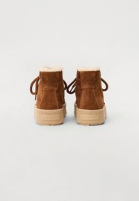 Bottines en daim marron avec un col en fourrure synthétique crème, lacets ronds et semelle beige texturée. Logo embossé à l'arrière.