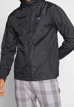 Mand iført en sort lynlåstrøje vindjakke og grå-hvide ternede bukser, med hænder placeret nær taljen.