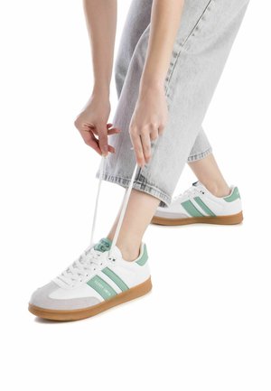 Zapatillas blancas con detalles verdes, una parte superior texturizada y una suela de goma. La persona está atando los cordones mientras lleva unos pantalones vaqueros claros.