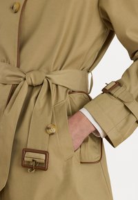 Trench-coat beige avec une taille ceinturée, des accents marron, un grand bouton et des poches avec un passepoil contrastant. Texture de tissu lisse.