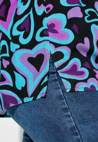 Buntes schwarzes Oberteil mit großen, mehrfarbigen Herzen in Blau- und Lilatönen. Ein Seitenschlitz ergänzt den lockeren Schnitt.