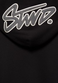 Black fabric with a large stitched gray and white signature-style text reading "StwiD." on the upper part.