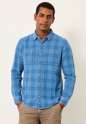 Man standing against white background wearing blue plaid button-up shirt and beige pants with hands in pockets.