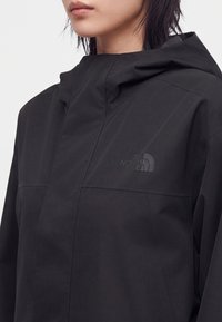 Personne portant une veste imperméable noire à capuche avec un col haut et un logo North Face discret sur la poitrine, visible des épaules vers le haut.