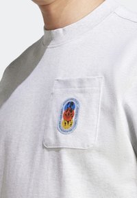 Detailní záběr na světle šedou mikinu se záševkem na prsou, která má vícerozměrnou nášivku loga Adidas Originals.