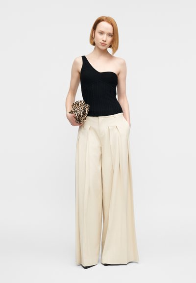 Haut noir côtelé à une épaule associé à un pantalon beige plissé, taille haute et jambes larges. Le modèle tient une pochette à motif léopard.