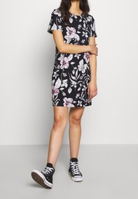 Květinové šaty s krátkým rukávem, tmavé pozadí, bílé a růžové květy, a přiléhavý střih, spárovány s černými vysokými teniskami.