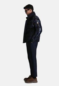 Doudoune marine à col montant et plusieurs poches, portée par-dessus un pantalon noir taillé. Des chaussures en daim marron complètent l'ensemble.