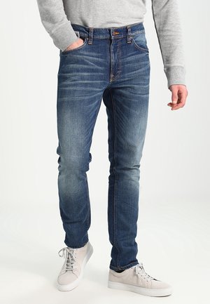 Homme portant un jean slim bleu délavé, un pull gris clair et des baskets blanches, debout avec une main dans la poche.