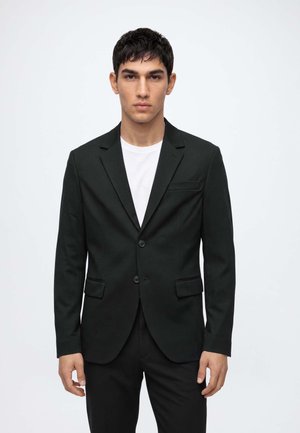 Un homme se tient face à l'objectif, portant une veste de costume noire sur une chemise blanche, sur un fond clair uni.