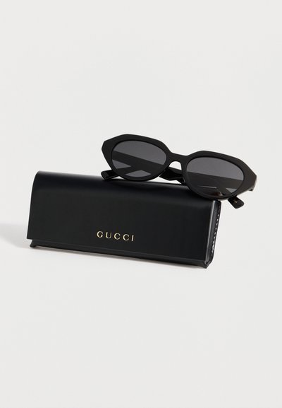 Lunettes de soleil noires angulaires posées sur un étui Gucci noir avec un logo doré sur un fond blanc uni.