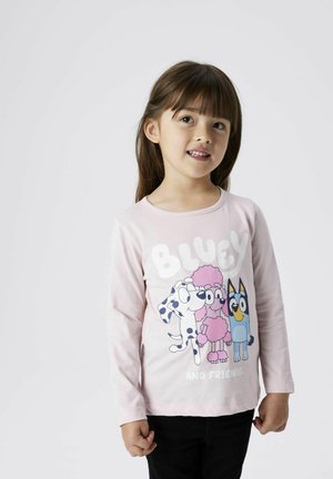 T-shirt rose à manches longues présentant des personnages de dessin animé avec le texte "BLUEY ET SES AMIS" en lettres blanches en gras. Pantalons noirs portés en dessous.