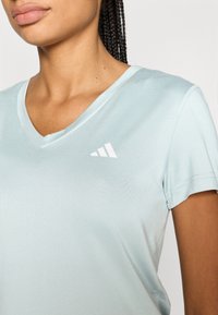 Αθλητική μπλούζα με V λαιμόκοψη σε ανοιχτό γαλάζιο, κατασκευασμένη από λείο, ελαστικό ύφασμα, με λευκό λογότυπο Adidas στην αριστερή πλευρά του στήθο�υς.