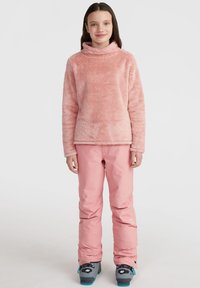 Rosa Fleecepullover mit hohem Kragen, kombiniert mit rosa Schneeanzug. Der Pullover hat eine weiche Textur und das Outfit beinhaltet Skischuhe.