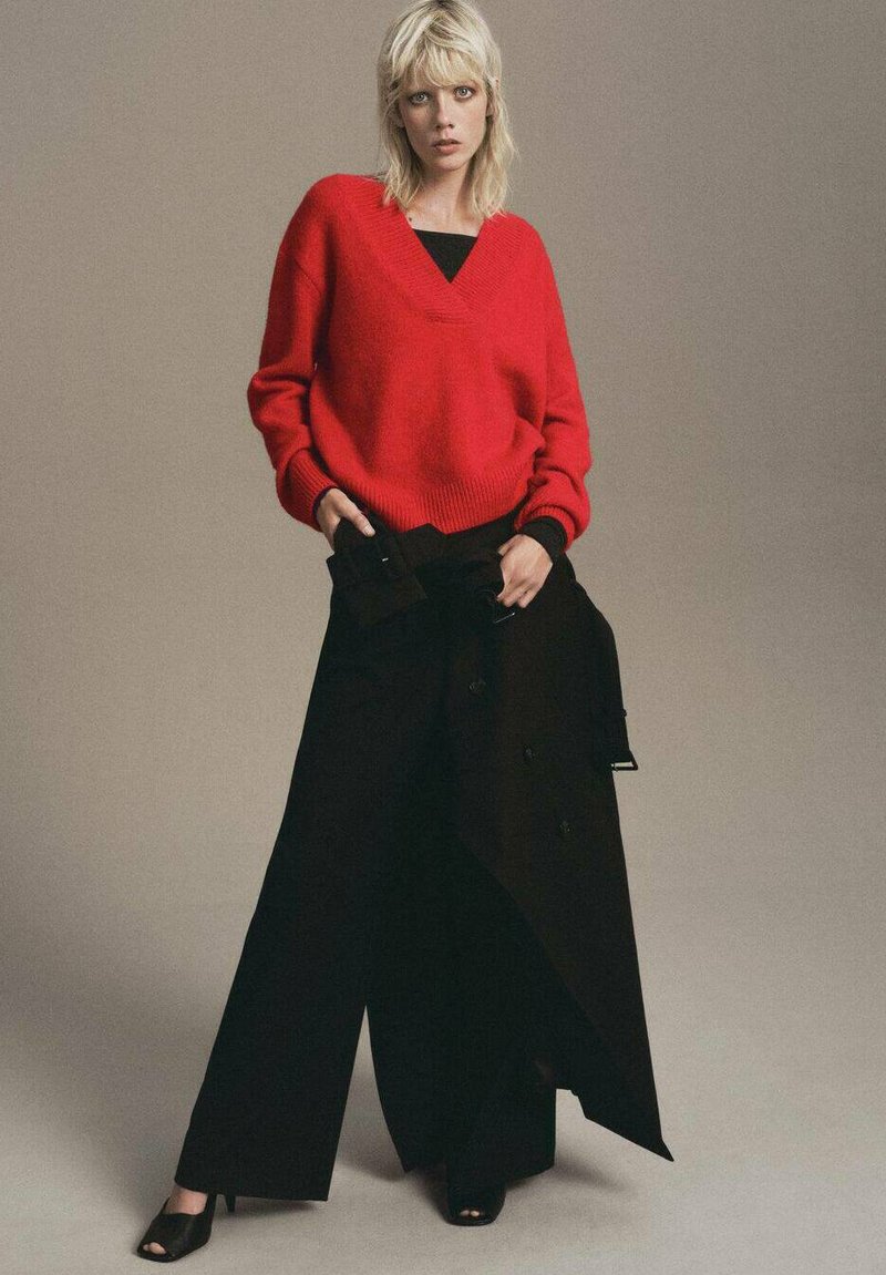 Pull en tricot rouge avec un col en V et des poignets côtelés, associé à un pantalon large noir avec une ceinture latérale et des boutons. Chaussures à talons blocs noires.