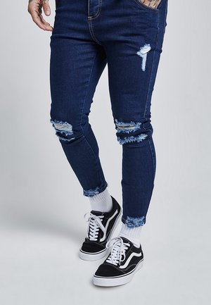 Ben iført mørkeblå slidte skinny jeans, hvide sokker og sort-hvide snørebåndssneakers på en ensfarvet lys baggrund.