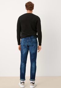 Hombre de pie de espaldas que lleva una camisa negra de manga larga, jeans azules remangados, calcetines blancos y zapatillas beige y negras.