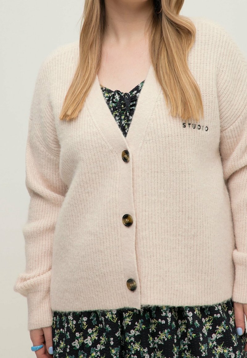 Žena na sobě má krémový pletený cardigan se třemi knoflíky přes černé květinové šaty, zobrazená je od pasu nahoru, včetně rukou s prsteny.