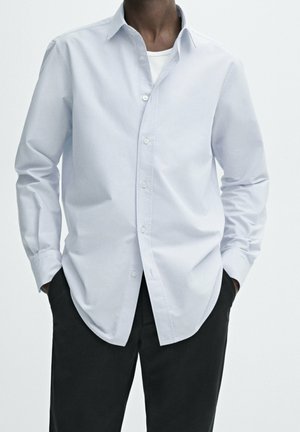 Man wearing light blue striped button-up shirt over white undershirt and black pants, hands in pockets, standing against plain background.