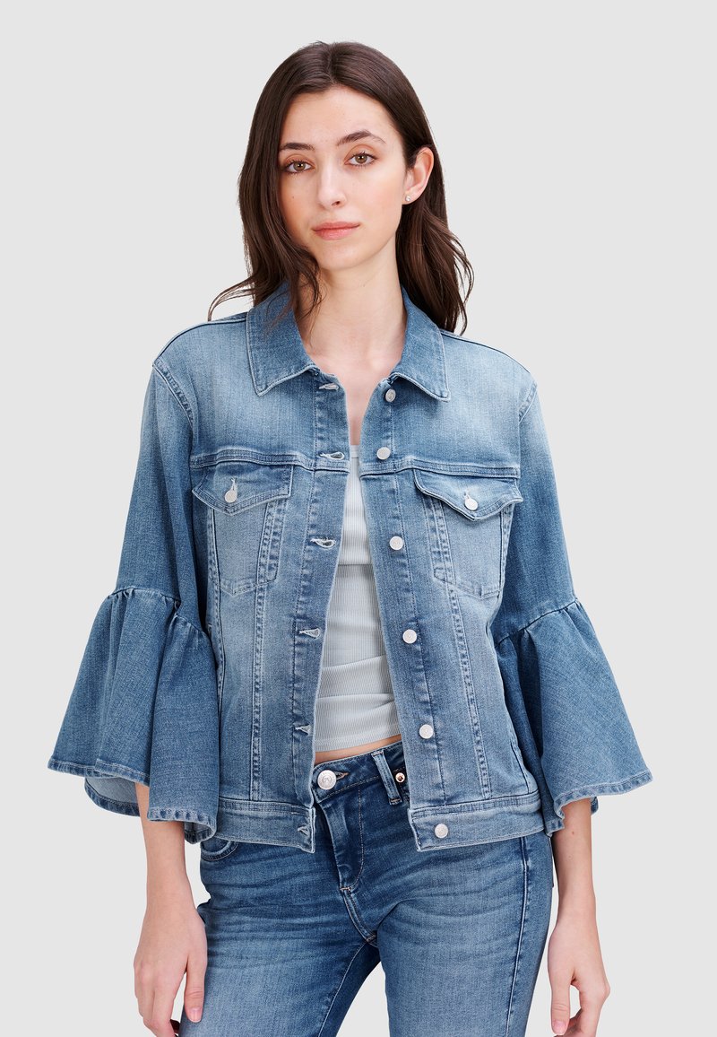 Hellblaue Jeansjacke mit ausladenden Ärmeln, vorderen Knöpfen, zwei Brusttaschen und fransigem Saum, kombiniert mit einem enganliegenden grauen Oberteil und Jeans.
