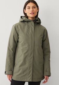 Donna che indossa una giacca impermeabile verde oliva con cappuccio, tasche frontali e polsini regolabili, in piedi davanti a uno sfondo chiaro e semplice.