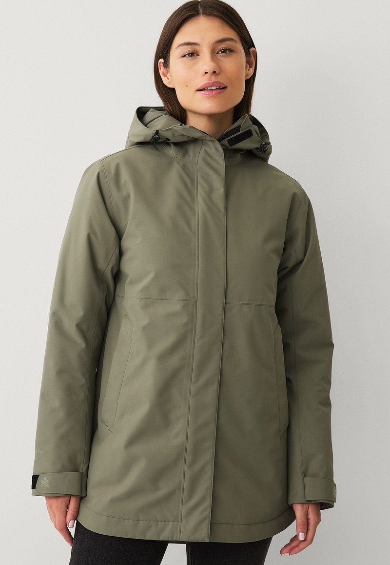 Donna che indossa una giacca impermeabile verde oliva con cappuccio, tasche frontali e polsini regolabili, in piedi davanti a uno sfondo chiaro e semplice.
