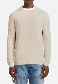 Maglione beige a costine con scollatura a girocollo e maniche lunghe, caratterizzato da un tessuto testurizzato e polsini aderenti, abbinato a pantaloni in denim blu.