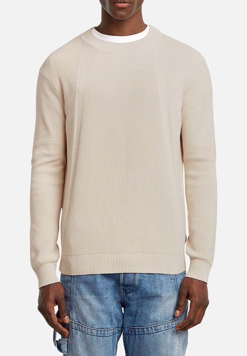 Maglione beige a costine con scollatura a girocollo e maniche lunghe, caratterizzato da un tessuto testurizzato e polsini aderenti, abbinato a pantaloni in denim blu.