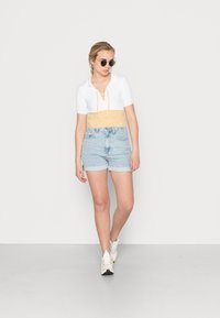 Top branco cropped com textura canelada e frente com atacadores, combinado com calções de ganga azul claro e ténis brancos. Os acessórios incluem óculos de sol redondos.