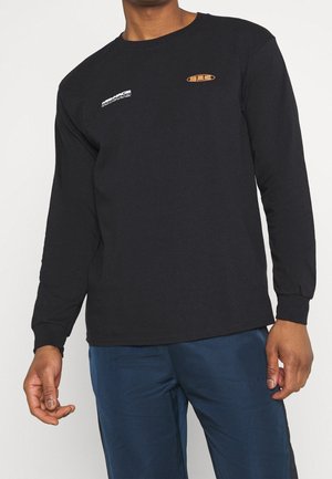 Personne portant un t-shirt noir à manches longues avec de petits logos texte blancs et orange sur la poitrine et un pantalon bleu.