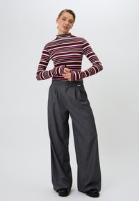 Gestreepte coltrui in bordeauxrood, roze en wit. Brede, grijs gestreepte broek met plooien en knoopdetails. Zwarte schoenen.