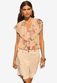 Blusa floreale in rosa chiaro con colletto con pizzo e chiusura a bottoni, abbinata a pantaloni corti beige con dettaglio a bottoni sul fronte.